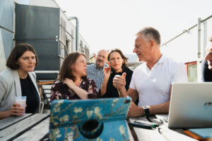 Man sieht mehrere Menschen im angeregten Gespräch miteinander. Sie wirken professionell, engagiert und fokussiert. Vor ihnen sieht man Stifte und Laptops. Sie scheinen sich auf einer Dachterasse oder etwas ähnlichem zu befinden.