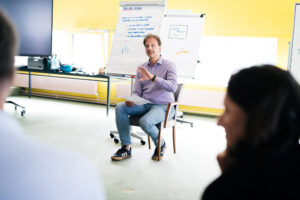 Ein Mann sitzt in einem Stuhl vor einer Gruppe von Teilnehmern und gestikuliert während eines Vortrags. Im Hintergrund sind Flipcharts mit Notizen und Zeichnungen zu sehen. Die Szene findet in einem hellen, freundlichen Seminarraum statt.