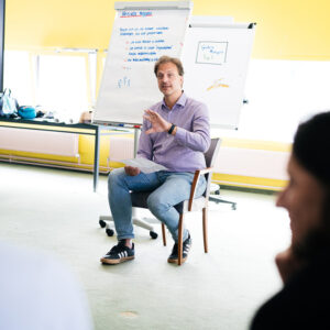 Ein Mann sitzt in einem Stuhl vor einer Gruppe von Teilnehmern und gestikuliert während eines Vortrags. Im Hintergrund sind Flipcharts mit Notizen und Zeichnungen zu sehen. Die Szene findet in einem hellen, freundlichen Seminarraum statt.