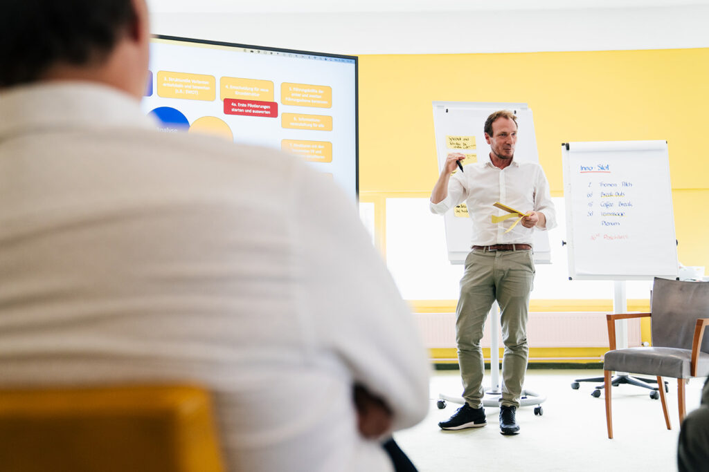 Ein Mann steht vor einem großen Bildschirm und präsentiert ein Konzept, während er auf die Inhalte zeigt. Im Hintergrund sind Flipcharts mit Notizen zu sehen, die auf eine interaktive Workshop-Situation hinweisen. Der Raum ist hell und in gelb gehalten, was eine freundliche Atmosphäre schafft.