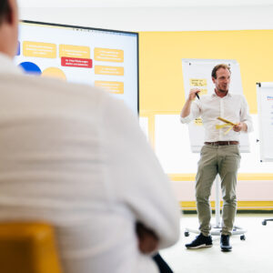 Ein Mann steht vor einem großen Bildschirm und präsentiert ein Konzept, während er auf die Inhalte zeigt. Im Hintergrund sind Flipcharts mit Notizen zu sehen, die auf eine interaktive Workshop-Situation hinweisen. Der Raum ist hell und in gelb gehalten, was eine freundliche Atmosphäre schafft.