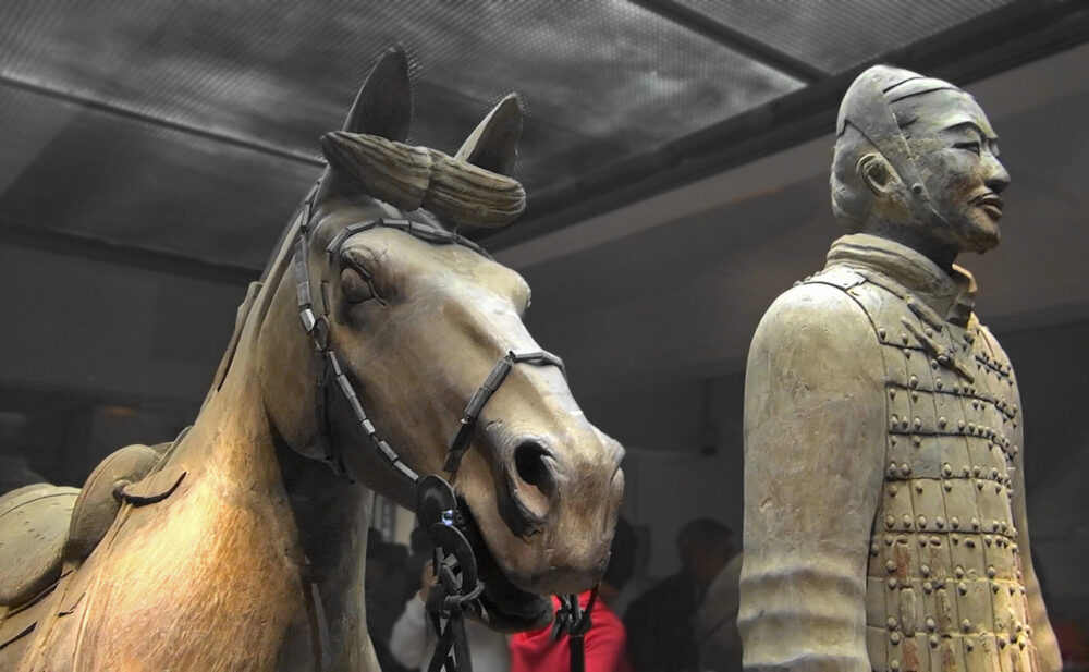 Nahaufnahme von zwei Statuen der Terrakotta-Armee: ein Pferd und ein Soldat. Das Pferd trägt ein Zaumzeug und hat ein detailliert ausgearbeitetes Gesicht. Der Soldat steht aufrecht daneben, gekleidet in eine Rüstung mit fein gearbeiteten Details. Beide Figuren sind Teil der berühmten Terrakotta-Armee aus China, die als Grabbeigabe für den ersten Kaiser von China, Qin Shi Huang, geschaffen wurde.