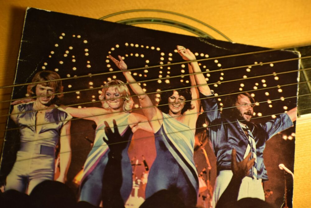 Foto einer Schallplattenhülle der schwedischen Popgruppe ABBA, auf der die vier Bandmitglieder Agnetha, Anni-Frid, Björn und Benny in glitzernden Bühnenoutfits zu sehen sind. Sie stehen Arm in Arm und winken dem Publikum zu, während im Hintergrund leuchtende Lichter die Bühne erhellen. Die Schallplattenhülle liegt auf einer Gitarre, deren Saiten im Vordergrund des Bildes sichtbar sind, was eine interessante Überlagerung der Texturen schafft.