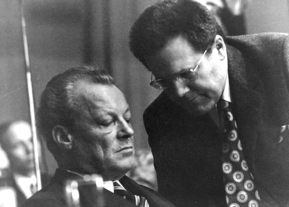 Schwarz-Weiß-Fotografie von Willy Brandt, dem ehemaligen Bundeskanzler der Bundesrepublik Deutschland, der nachdenklich und mit gesenktem Blick sitzt. Neben ihm steht Günter Guillaume, der ihm etwas ins Ohr flüstert. Guillaume war einer der persönlichen Referenten Brandts, später als DDR-Spion enttarnt. Das Bild fängt einen ernsten Moment ein und ist historisch bedeutsam, da die Enthüllung von Guillaumes Spionagetätigkeit zur sogenannten Guillaume-Affäre führte.